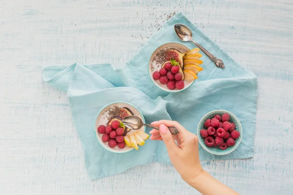 Taze meyve, chia tohum, ahududu ve sağlıklı vegan vejetaryen diyet kahvaltı için beyaz rustik ahşap arka plan üzerinde incir ile güler yüzlü kase. Sağlıklı gıda kavramı. Kadın eli bir kaşık tutan. Üstten görünüm