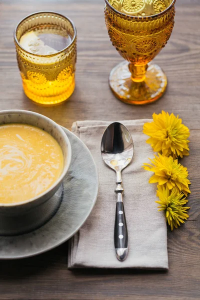 Tavolo rustico con zuppa di zucca, tovagliolo di lino, posate, piatti in ceramica, bicchieri gialli e fiori gialli — Foto Stock