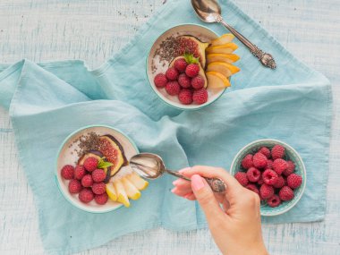 Taze meyve, chia tohum, ahududu ve sağlıklı vegan vejetaryen diyet kahvaltı için beyaz rustik ahşap arka plan üzerinde incir ile güler yüzlü kase. Sağlıklı gıda kavramı. El bir kaşık tutan. Ek yük