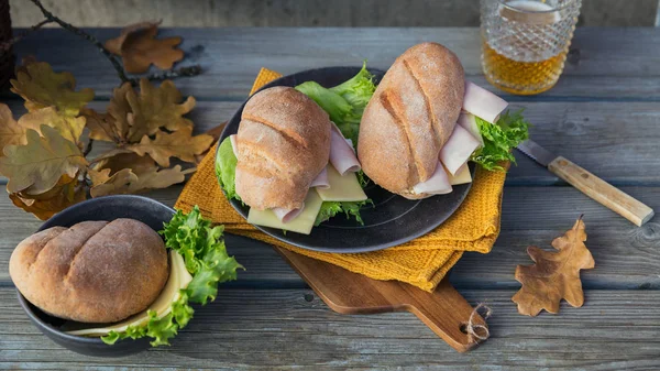 Two fresh ciabatta baguette sandwiches with ham, cheese, lettuce and a glass of beer on rustic wooden background. Outdoor autumn picnic lifestyle.