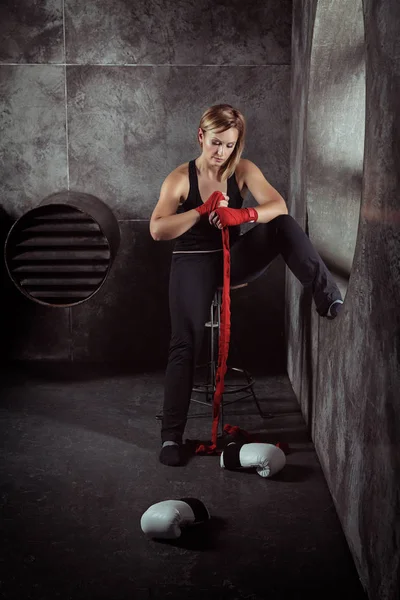 Sporting blonde woman in boxing bandages and boxing gloves around in dark studio. Woman is wrapping hands with red boxing wraps. Strong woman concept.