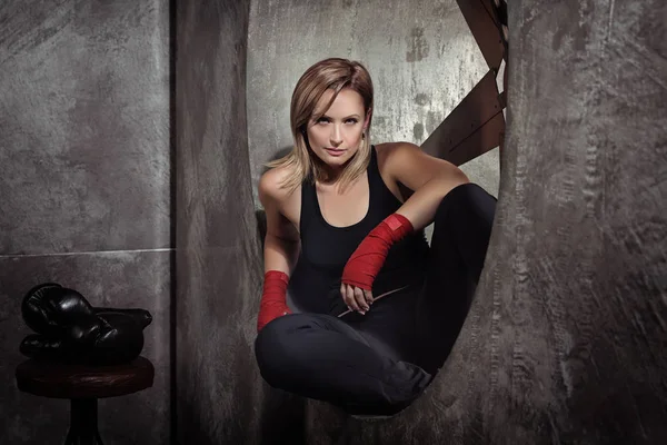 Sporting blonde woman in boxing bandages and boxing gloves around in dark studio looking at camera. Strong woman concept.