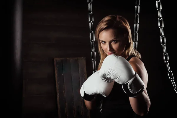 Sporting blonde woman in boxing gloves in dark studio looking at camera. Woman self defense concept.