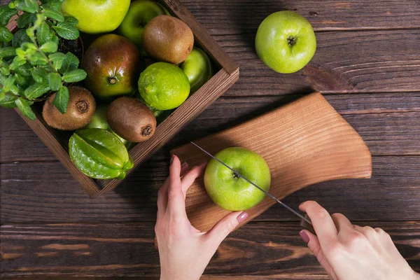 Woman cutting green apple on wooden cutting board. Fresh green fruits in a box on dark wooden background. Set of green fruits for healthy diet and detox: apple, lime, kiwi, mango, carambola and mint.
