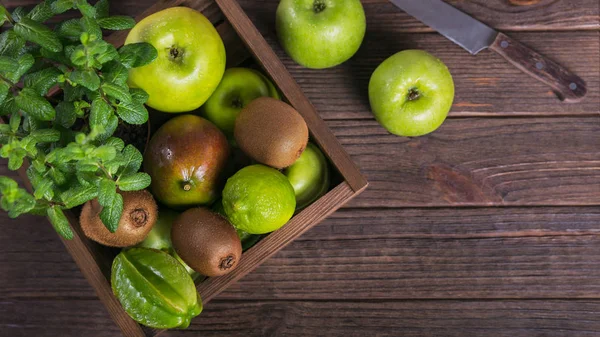 Healthy food background. Concept of healthy food. Fresh green fruits in box on dark wooden background. Set of green fruits for healthy diet and detox: apple, lime, kiwi, carambola and mint. Top view