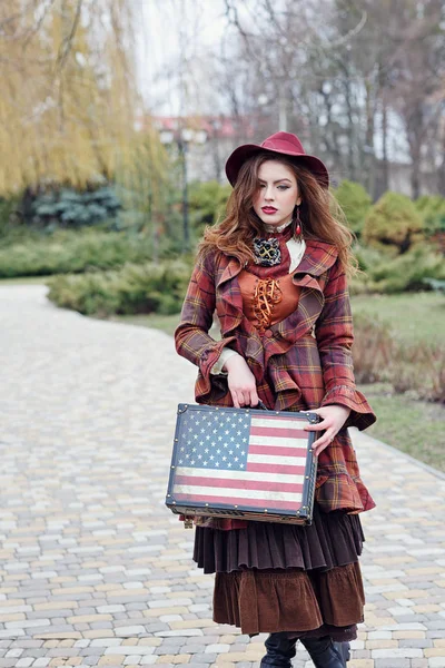 Beautiful woman in a vintage clothing of the Victorian era and elegant hat goes along the road with a suitcase in the American style