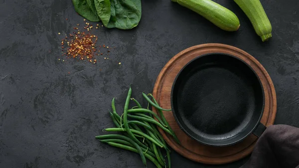 Draufsicht auf Zucchini, Spinatblätter, Gewürze und grüne Bohnen um leere Kochpfanne auf dunklem Tisch. Zutat zum Kochen gesundes Frühstück Attrappe. — Stockfoto