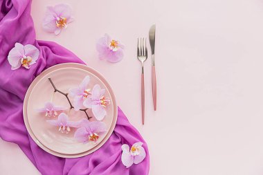 Açık pembe arkaplanda çiçek ve masa ayarları. Pembe seramik tabaklar, çatal bıçak takımı ve perple orkide çiçekleri. Tatil modern masa dekorasyonu. Romantik yemek..