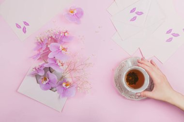 Sabah çayı, mor orkide çiçekleriyle dolu pembe bir mektup çantası ve açık pembe arka planda boş bir zarf. Kadın eli bir fincan çay tutuyor. Güzel hediye konsepti