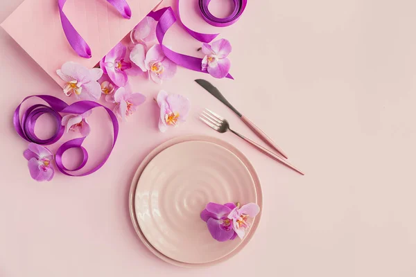 Flower and table settings overhead composition on light pink background. Pink ceramic plates, cutlery, gift bag with purple ribbons and orchid flowers. Holiday modern table decoration. Romantic dinner