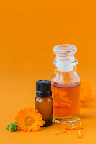 Calendula producten. Flessen cosmetische, aromatische of etherische olie en verse calendula bloemen op oranje achtergrond. Aromatherapie, spa, wellness concept — Stockfoto