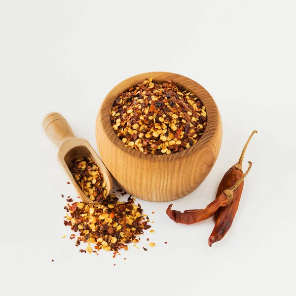 Wooden bowl and scoop full of crushed red cayenne pepper, dried chili flakes and seeds isolated on white background. Homemade spices ingredients for cooking.