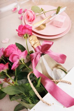 A stylish dinner arrangement with pink plates, golden cutlery, fresh roses, a champagne bottle with a bow, and delicate decor.