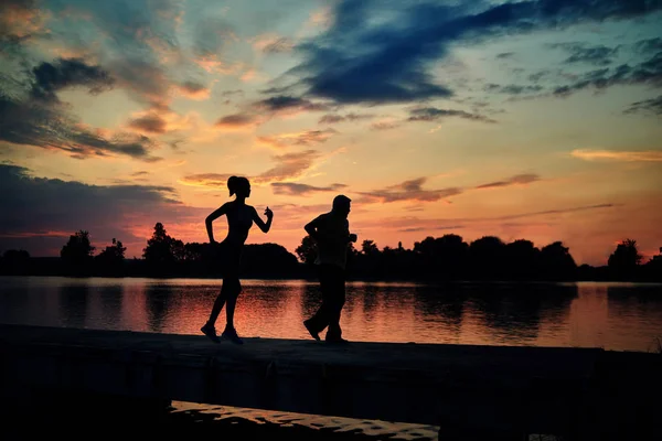 Ciemne sylwetki scamper biegaczy podczas zachodu słońca w pobliżu jeziora. — Zdjęcie stockowe