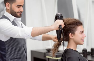 Arap hairstyler gülümseyen kadın istemcileri kıvırcık saç kurutma yan. Büyük ayna yansıması bulanık. Plastik fırça ve saç kurutma makinesi kullanarak. Beyaz gömlek, gri yelek giyiyor. Şık beatysalon.