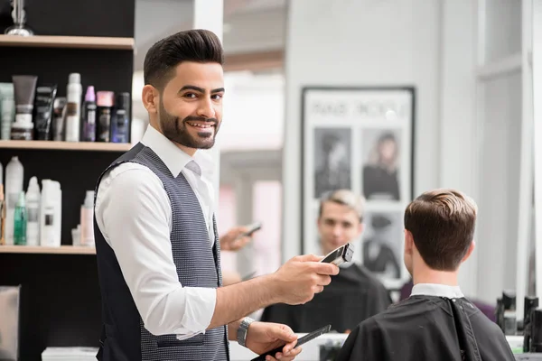 Улыбающийся парикмахер делает прическу для молодого клиента в парикмахерской . — стоковое фото