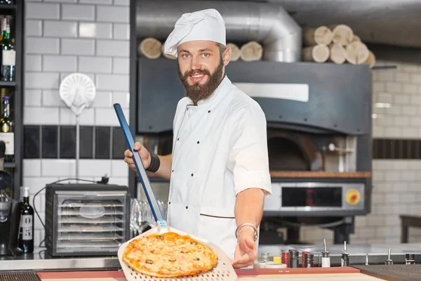 Vista frontale del fornaio che indossa la tunica degli chef e tiene la pizza sulla pala metallica . — Foto Stock
