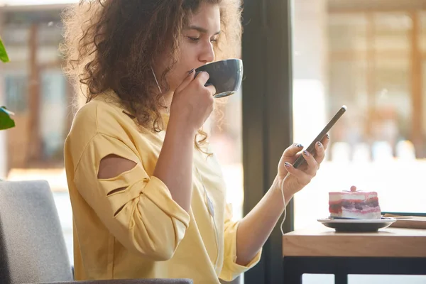 Chica rizada relajante beber café en la cafetería, utilizando el teléfono inteligente . — Foto de Stock