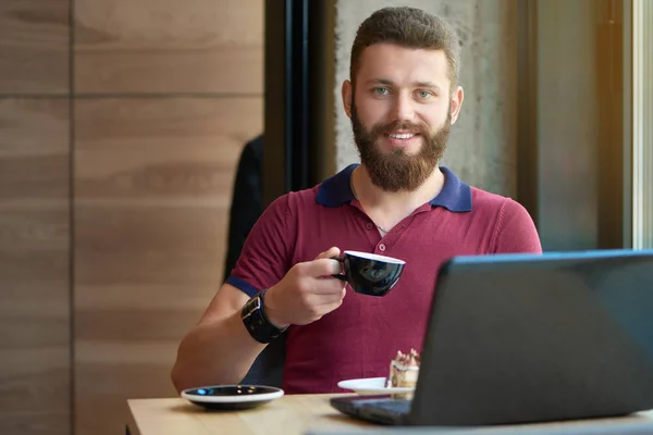 Sorrindo hipster mantendo xícara de café, trabalhando com laptop no café . — Fotografia de Stock