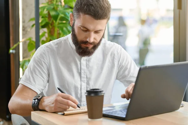 Homem barbudo escrevendo notas, trabalhando com laptop sentado perto da janela . — Fotografia de Stock