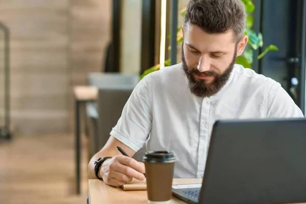 Bearder hipster escrever notas enquanto trabalhava com laptop, beber café . — Fotografia de Stock