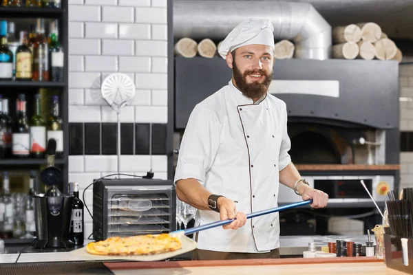 Panettiere sorridente indossa tunica bianca mantenendo lunga pala metallica con pizza . — Foto Stock