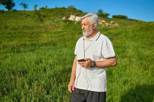 Vanha mies pitää kännykkää, seisoo kentällä katsoen sivusta . — kuvapankkivalokuva