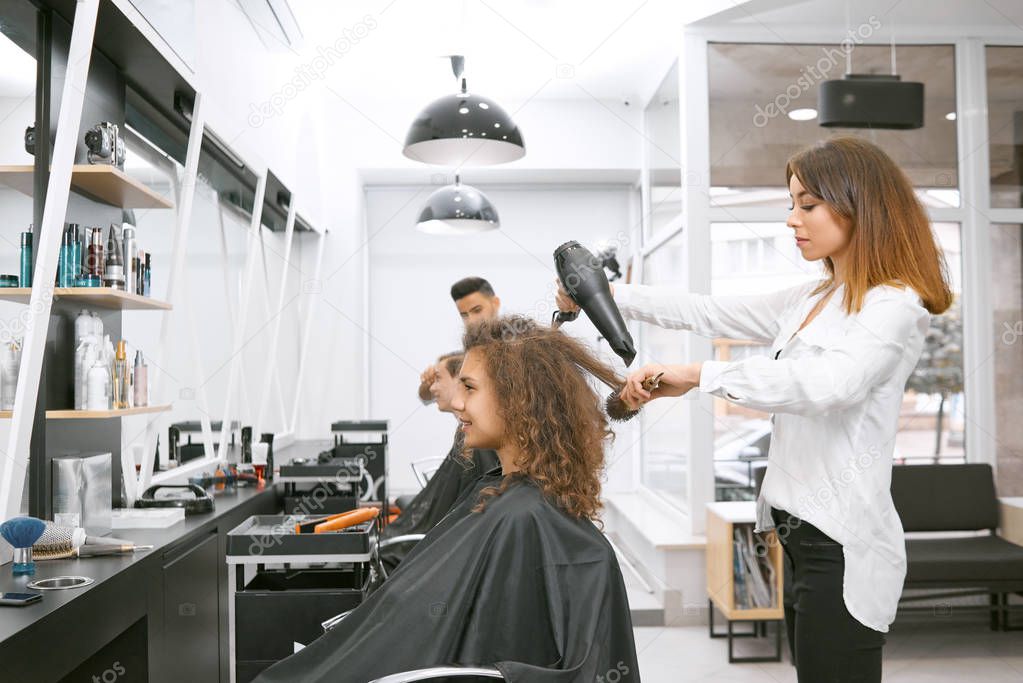 Female hairstylist drying curly girls hair using big plastic brush.