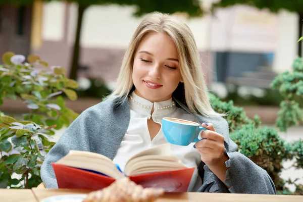 Linda chica rubia leyendo un libro sentado en cafés al aire libre salón . — Foto de Stock