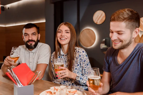 Två män och kvinna hålla glas öl och sitter i pizzeria. — Stockfoto