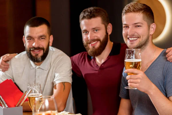 Três homens bonitos sorrindo, olhando para a câmera e segurando um copo de cerveja . — Fotografia de Stock