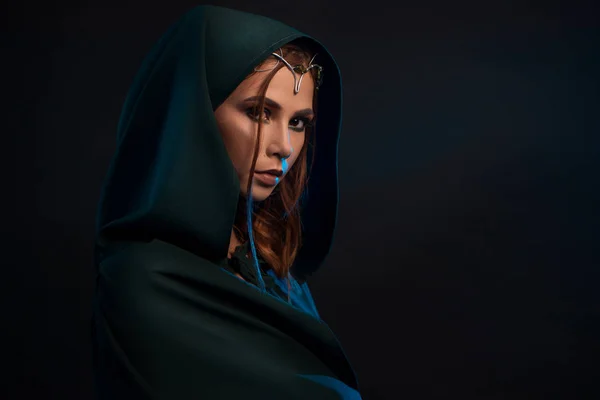 Beatiful elf princess wearing dark green cape looking at camera from darkness. — Stock Photo, Image