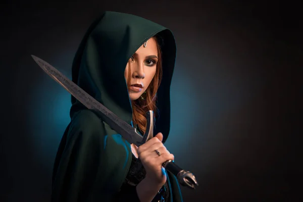 Mysterious young woman keeping sharp knife, wearing green cape with a hood. — Stock Photo, Image