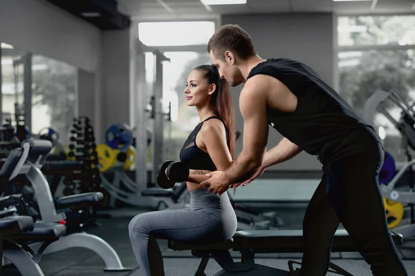 Handsome personal instructor helping his female client to training with dumbbells by supporting her muscular hand. Sports girl with long hair, laid in tail, workout in sexy sports uniform in dark gym.