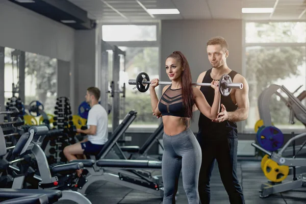 Male personal trainer supporting woman working with heavy barbell at gym.