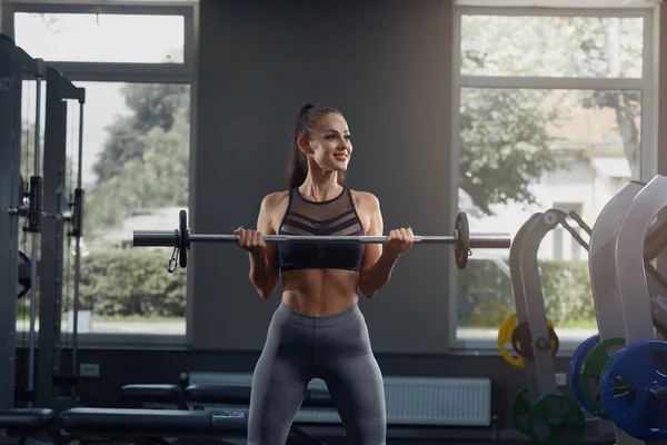 Heißes junges Mädchen im Fitnessstudio duckt sich mit Langhantel gegen Fitnessgeräte. — Stockfoto