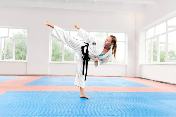 Sporty karate woman in white kimono with black belt against big window standing in karate position at fight class. Female fighter with blue eyes and braided hair improving technique of fight.