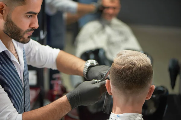 Handsome barber in black gloves cutting haircut of man using scissors.