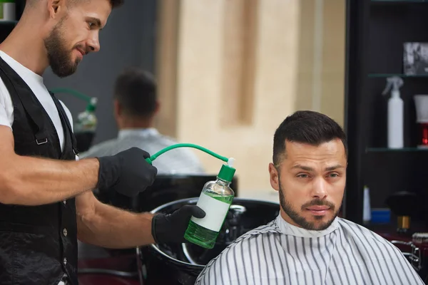 Barber in black gloves sprinkling green water on handsome man.