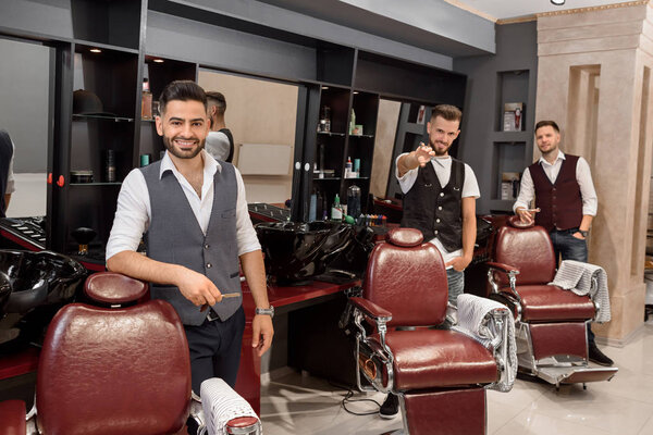 Three handsome barbers posing in barbershop near hairdresser chairs.
