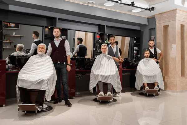 Barbiers debout près des clients couverts de capes et assis dans des chaises de coiffeur . — Photo