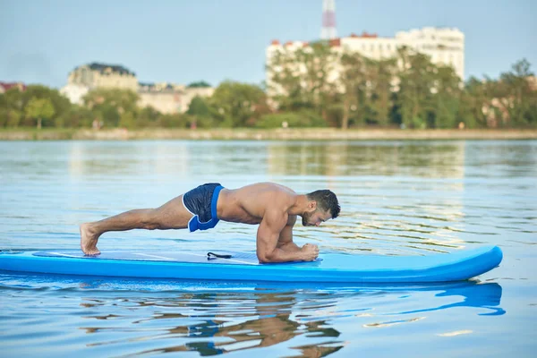 Hombre guapo haciendo ejercicio de tablón en el tablero de paddle en el lago de la ciudad . — Foto de Stock