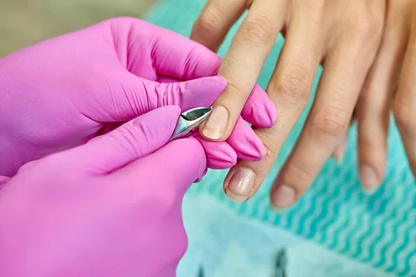 Closeup of process of removing cuticle. Hands of professional manicurist in protective gloves keeping scissors and cutting cuticle in salon. Concept of doing manicure and fingernails cleaning.