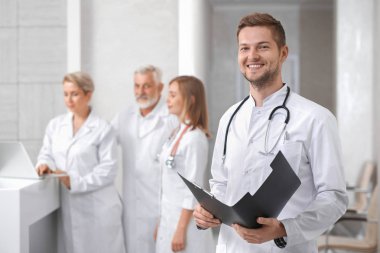 Bir profesyonel, özel hastane sağlık personeli. Yakışıklı erkek doktor ayakta, kameraya bakıyor ve gülümsüyor. Klasör holding ve boynunda steteskop sahip bir adam. Grup arkasında poz hekimlerin.