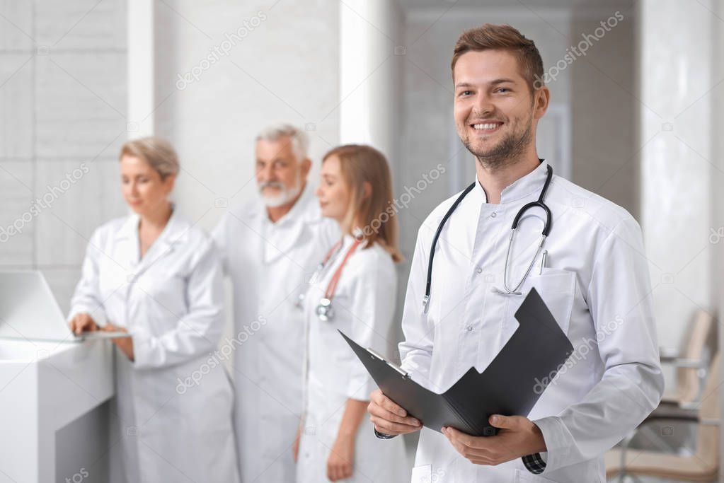 Medical staff of professional, private hospital. Handsome male doctor standing, looking at camera and smiling. Man holding folder and having stethoscope on neck. Group of physicians posing behind.