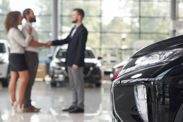 Close up of black car headlight. Couple making deal with car dealer behind. Man and manager of car showroom shaking hands, standing opposite. Purchase of vehicle.