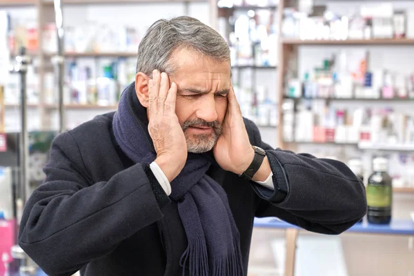 Ο άνθρωπος έχοντας πονοκέφαλος, αρπάζοντας το κεφάλι, στέκεται στο φαρμακείο. — Φωτογραφία Αρχείου