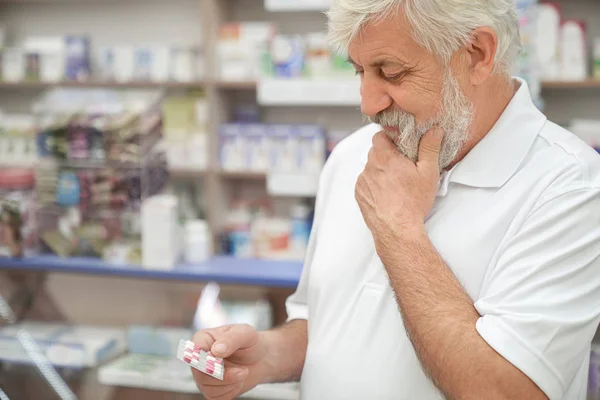 Elderly man thinking, holding pills in pharmacy. Stock Photo
