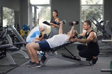 Modrn spor salonunda egzersiz kişiye kadar uygun. Dambıl ile elele güçlü adam bankta, yalan. Kişisel antrenör onu destekleyen. Kadın koç çalışma dışarı ile yaşlı adam yardım.