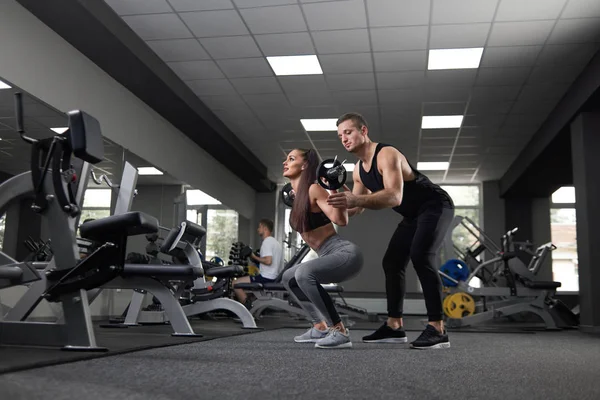 Brunette doen squats met barbell, trainer ondersteuning. — Stockfoto